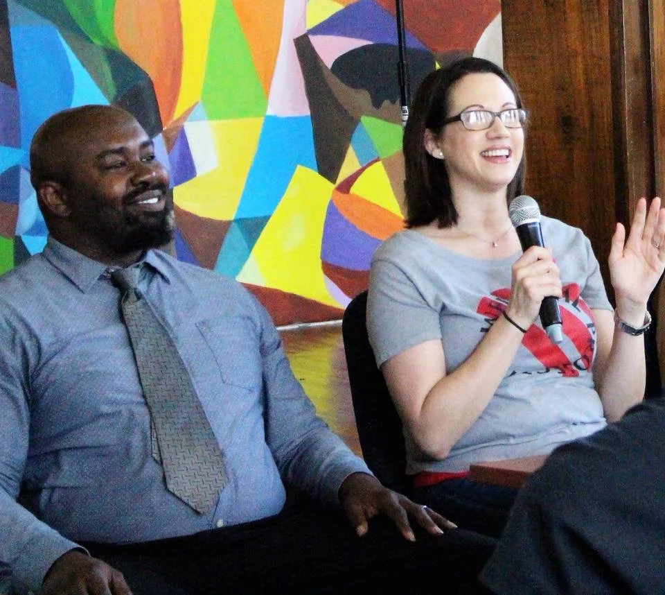 A Black man and a pale woman sitting at a table, both smiling, with a colorful abstract mural in the background. The woman is holding a microphone and speaking to an audience.
