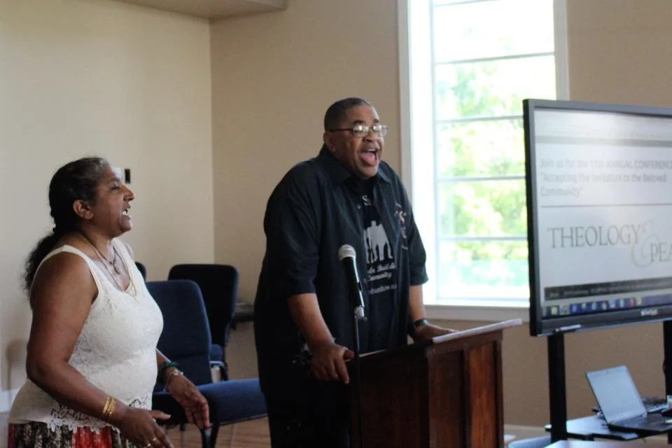 Two people are laughing and smiling in a room during a presentation or speech. One person is standing behind a wooden podium, and the other is standing next to them. There is a large screen displaying a presentation in the background, and a window le