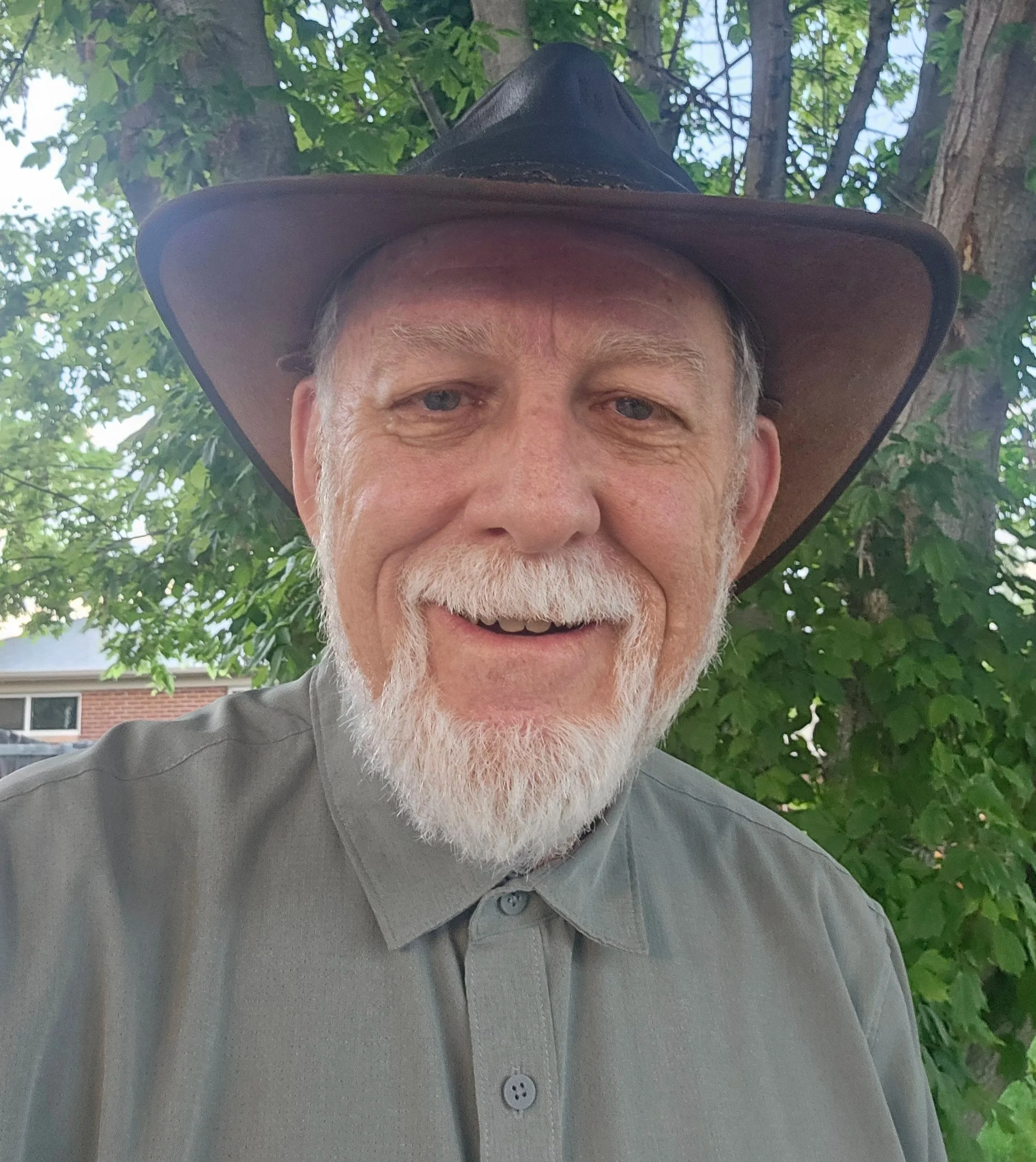 Close-up of an elderly man wearing a wide-brimmed brown hat and a gray collared shirt, outdoors with green trees and a building in the background.