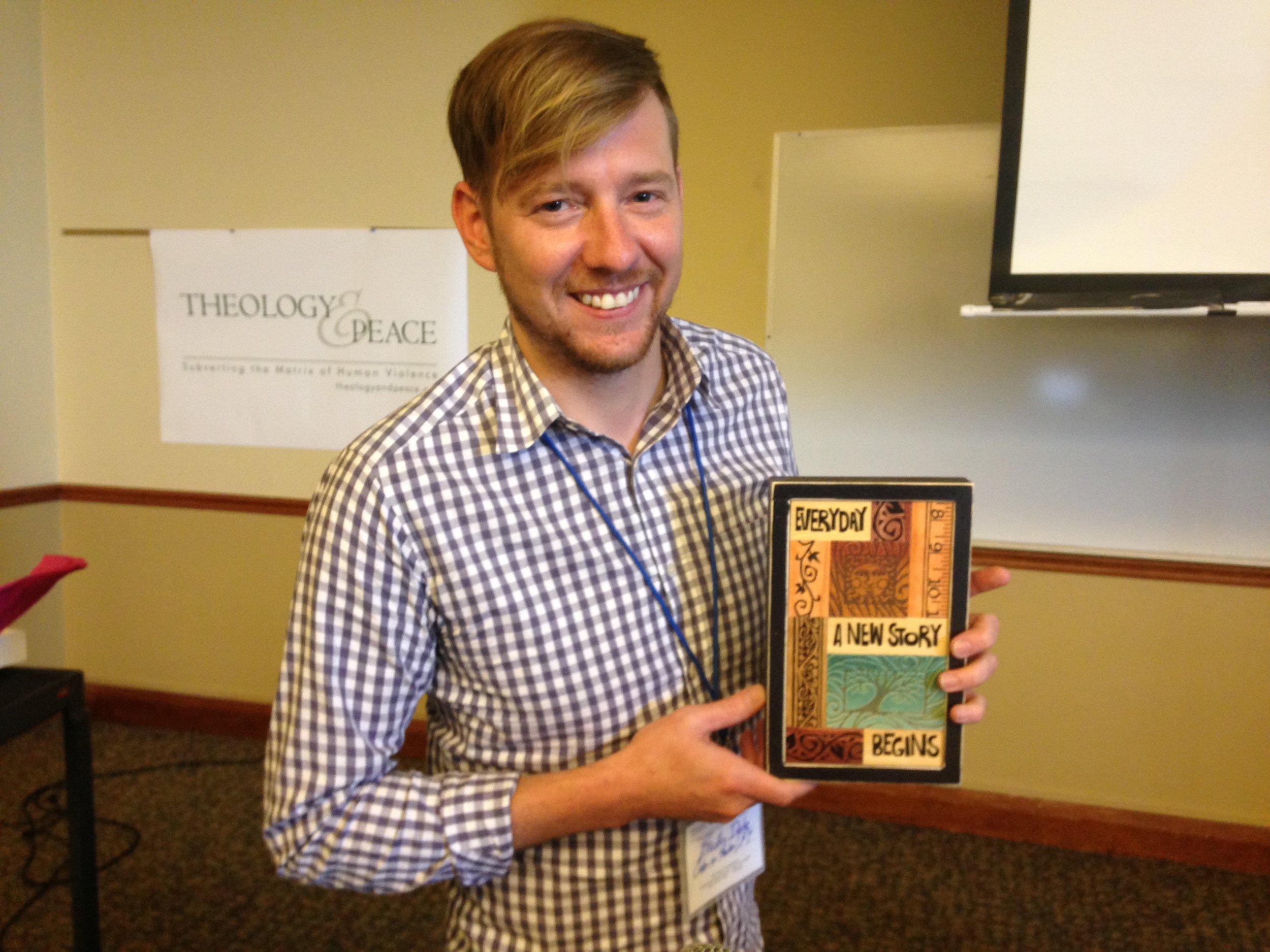 A smiling young man with light brown hair wearing a checkered shirt and a lanyard, holding a colorful book titled "Everyday A New Story Begins," standing in a conference room with a poster of the Theology & Peace logo in the background.