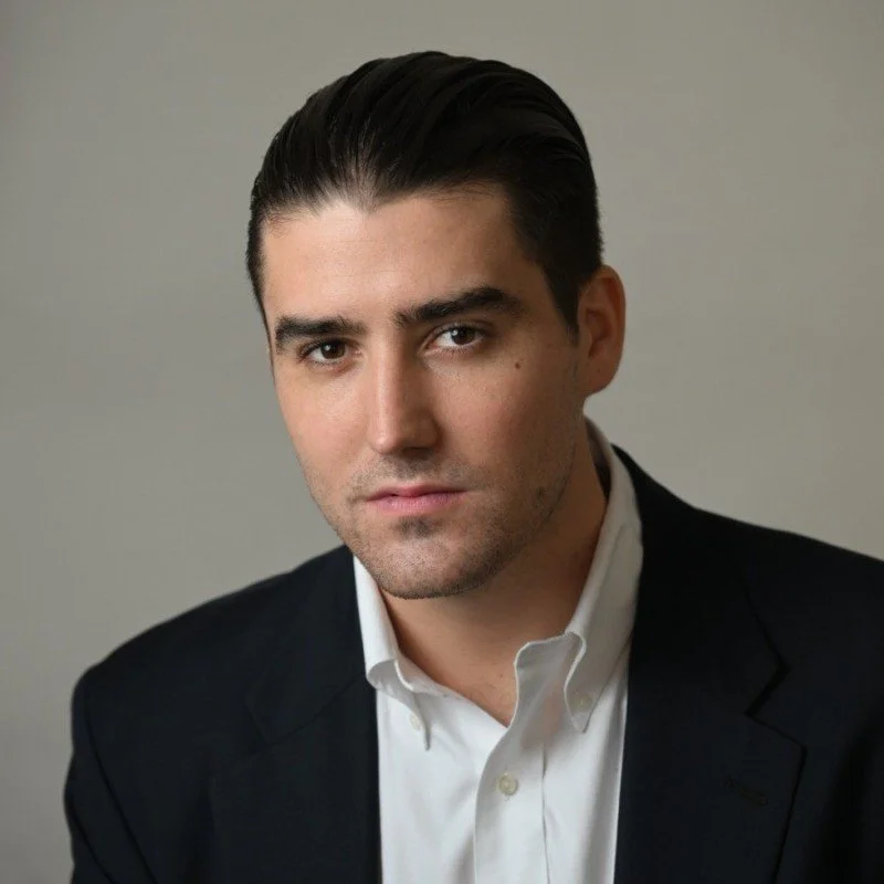 A young man with dark, neatly slicked-back hair, wearing a black blazer and white shirt, looking directly at the camera with a serious expression.