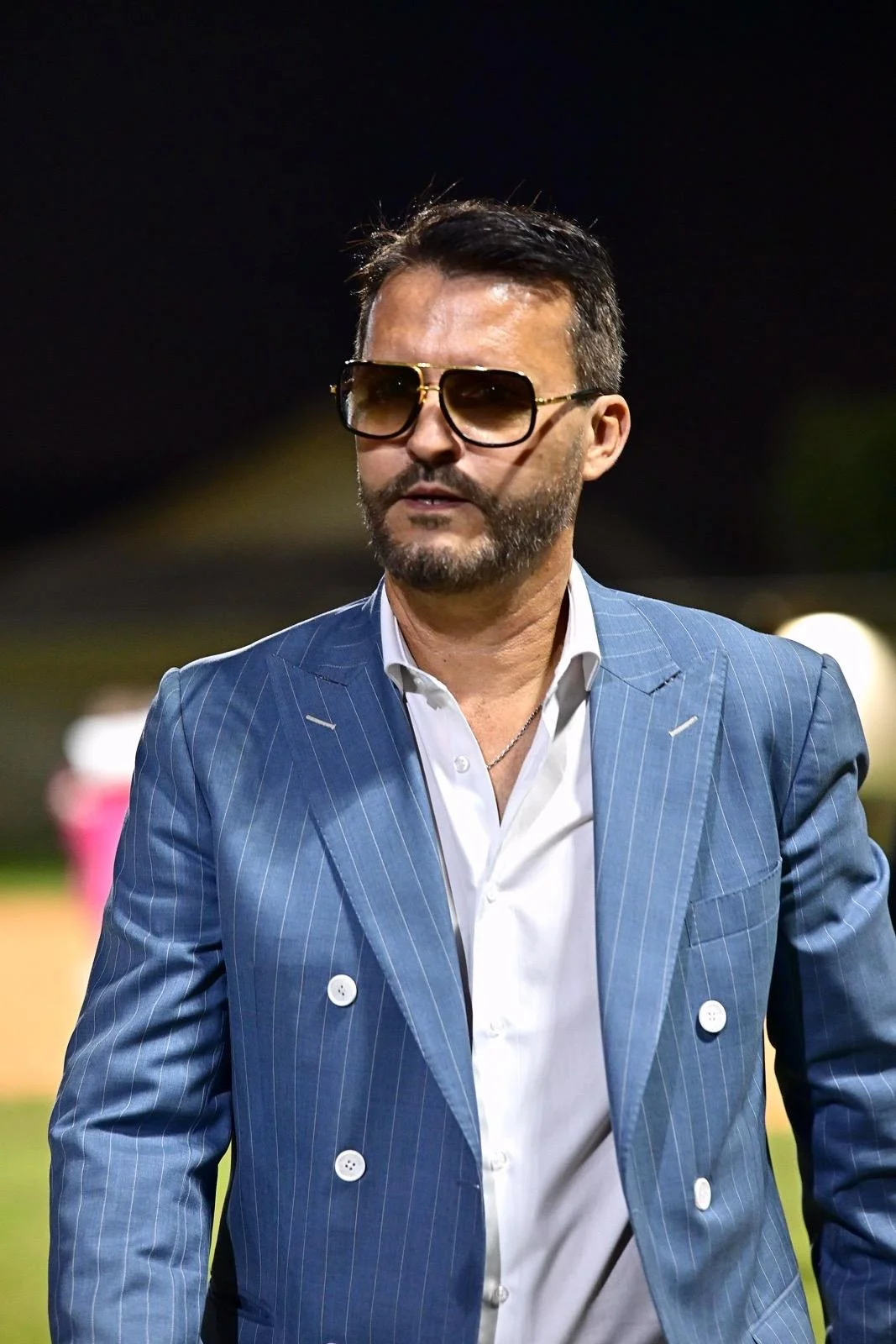 A man wearing a blue pinstripe suit, sunglasses, and a white shirt, standing outdoors at night.