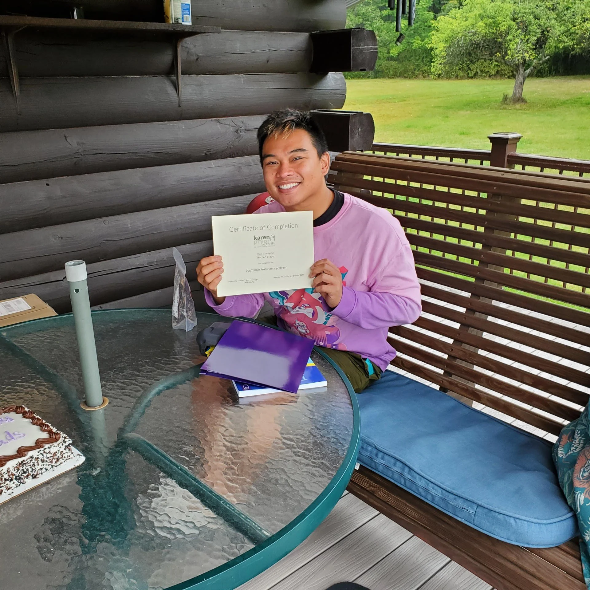 A young man sitting at an outdoor table on a porch, holding a certificate of completion and smiling. The table has a cake, books, and a water bottle on it. The porch has wooden logs as walls and a wooden bench with cushions. There is a grassy yard with trees in the background.