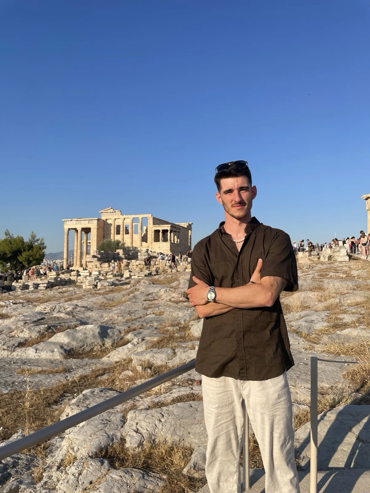 Ein junger Mann steht vor den antiken Ruinen der Akropolis in Athen, Griechenland, bei Sonnenschein und klarem blauen Himmel, mit Touristen im Hintergrund.