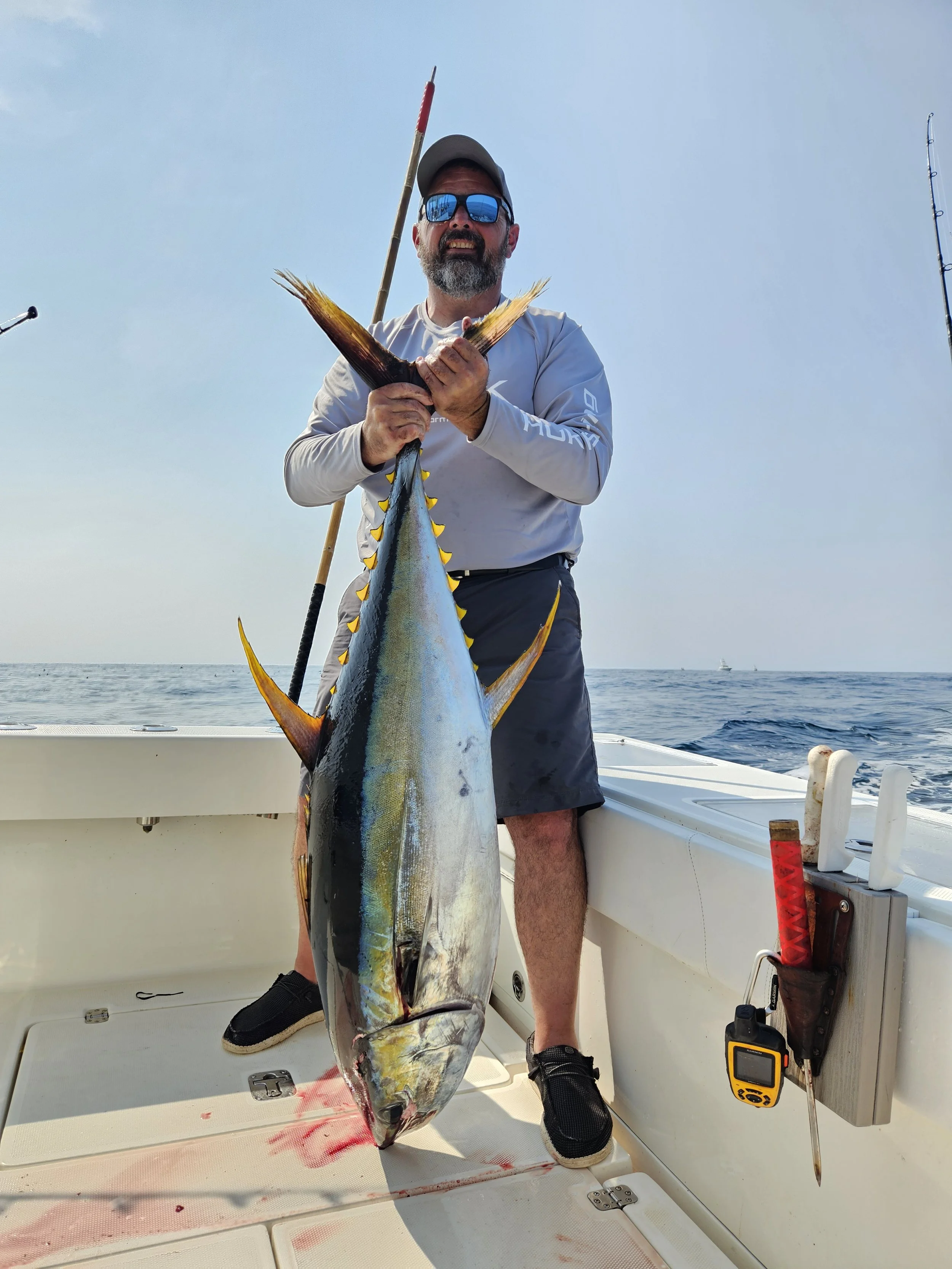 Man on a boat holding a large yellowfin tuna, fishing rods and a GPS device nearby, the sky is clear and ocean in the background. The charter fishing boat with Sea Farmer Charters