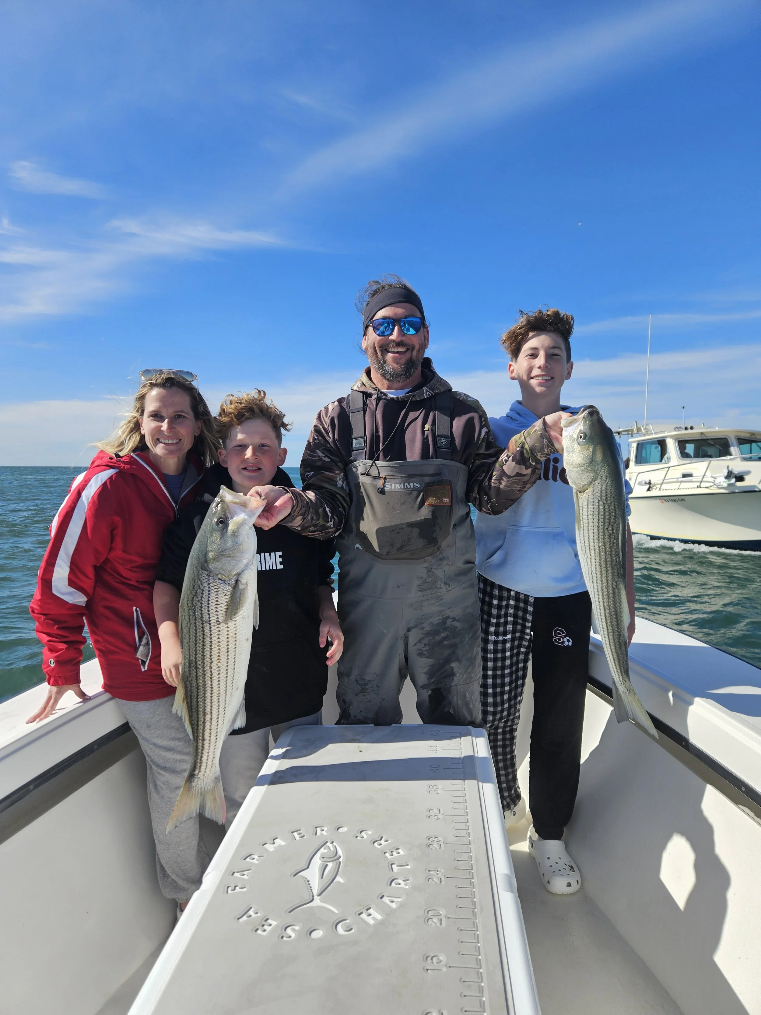 Family of four on a boat, holding caught fish, with a clear blue sky and a yacht in the background. Sea Farmer Charters