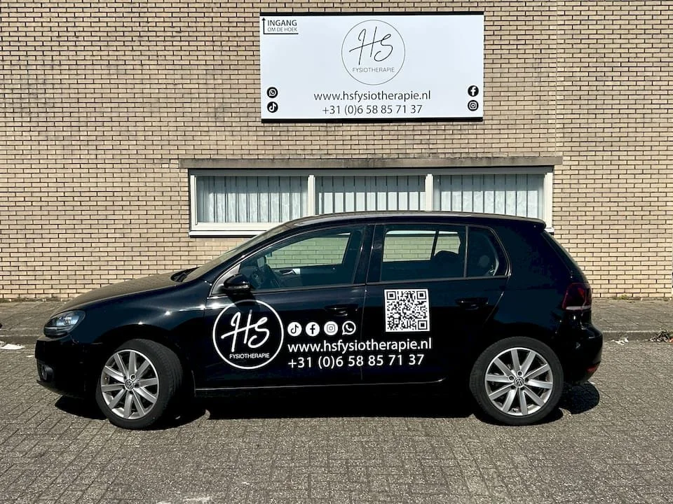Een zwarte auto met reclame voor een fysiotherapiepraktijk op de deur, geparkeerd langs een bakstenen muur met een groot wit bord erboven dat ook de praktijk advertentie bevat.