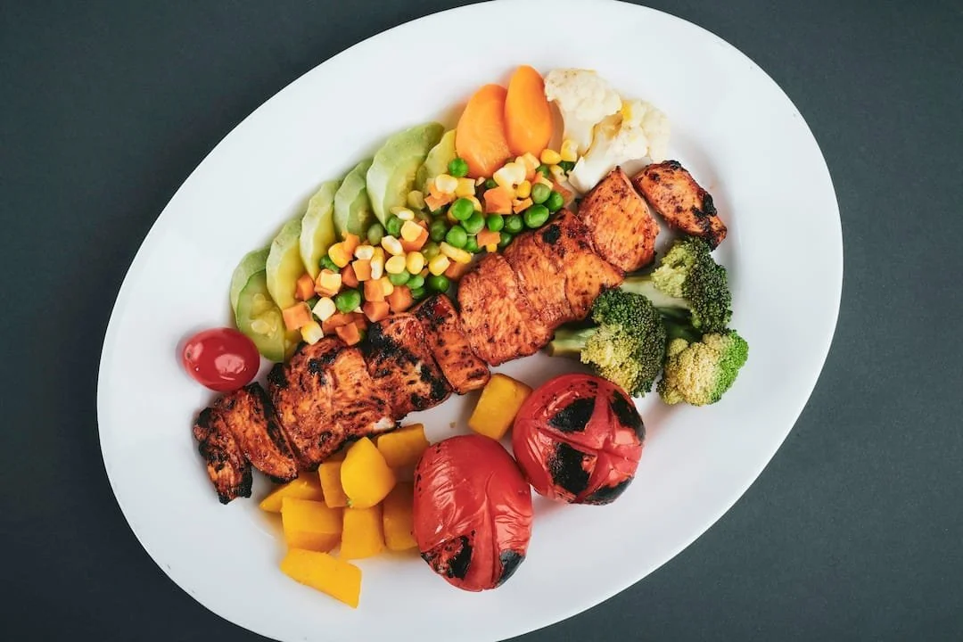 "Gegrilde kipspies met avocado en diverse groenten op een wit bord, van bovenaf gefotografeerd."