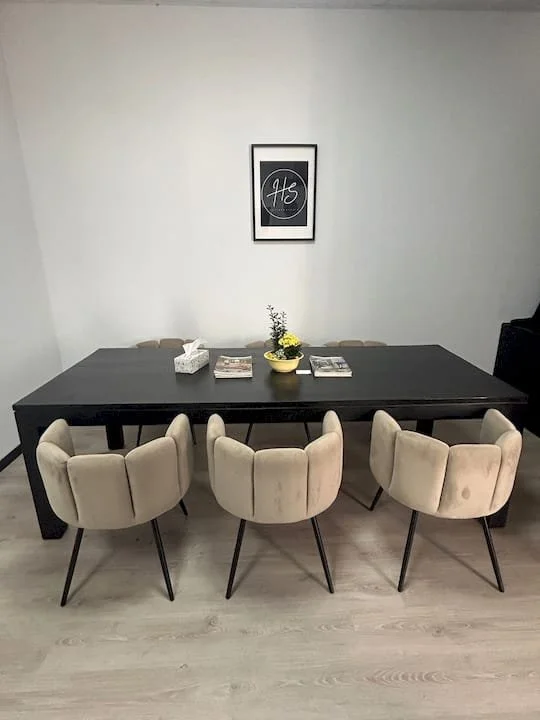 Eettafel met zes beige stoelen, een bloemstuk en boeken, tegen een witte muur met een zwart/wit ingelijste poster met de tekst 'He'.