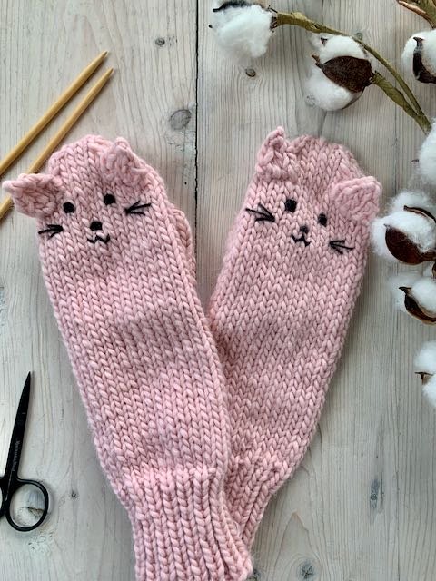 Two pink hand knitted mittens with ears and face of a cat, criss-crossed on a white wood table