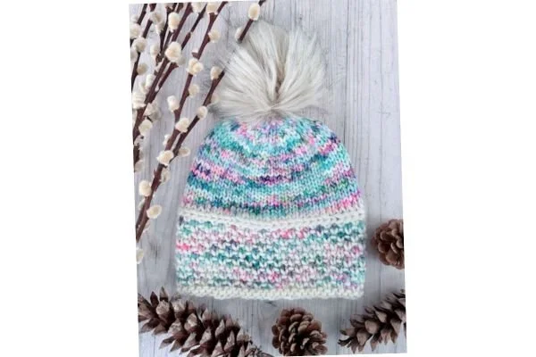 A hand knit hat with a brown pompom on a light wood table with pine cones