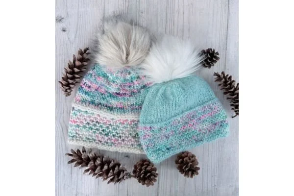 A pair of hand knits hats with pompoms on a light wood table with pine cones