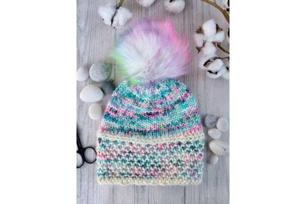 A hand knit hat with a rainbow pompom on a light wood table with pine cones