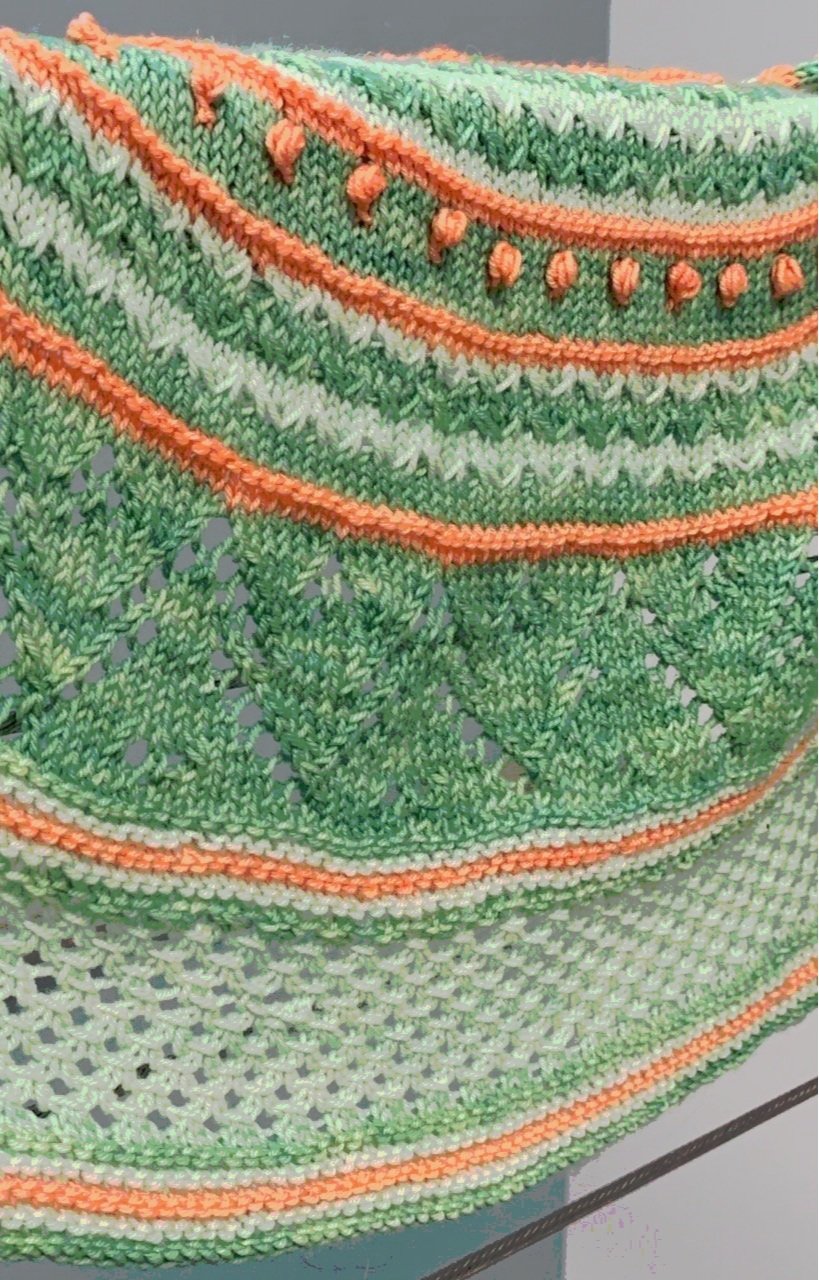 A hand knit crescent-shaped shawl in two shades of green plus orange