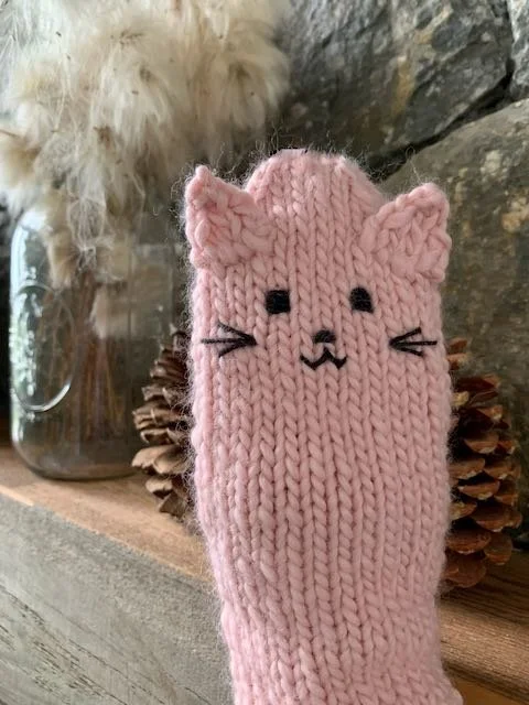 A pink hand knitted mitten with ears and face of a cat