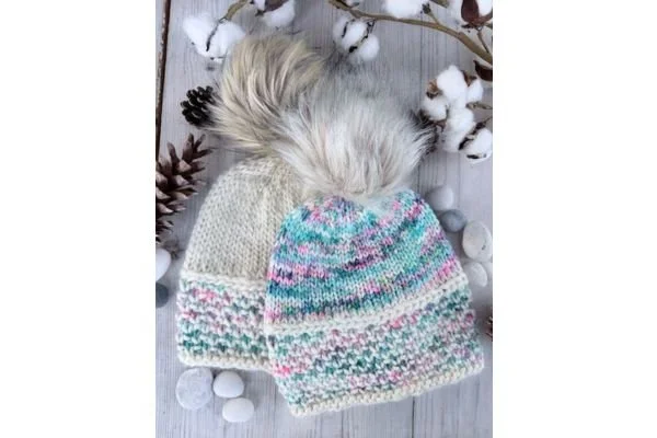 A pair of hand knit hats on a light wood table with pine cones