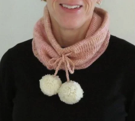 A smiling woman models a pink neck warmer with white pompoms