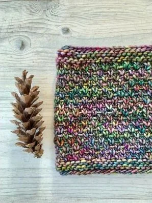 A colorful hand knit neck warmer on a light wood table alongside a pine cone
