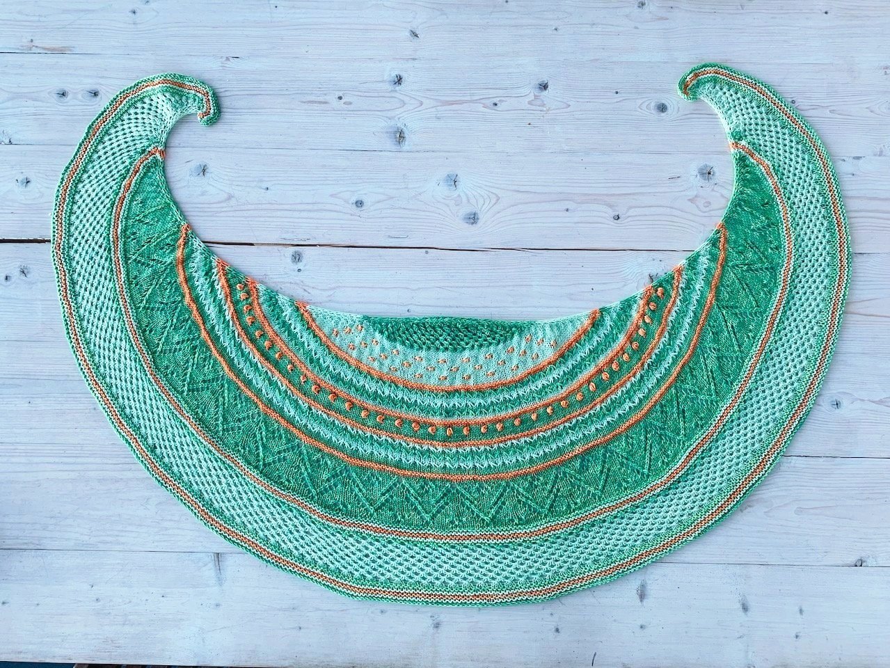 A hand knit crescent-shaped shawl in two shades of green plus orange