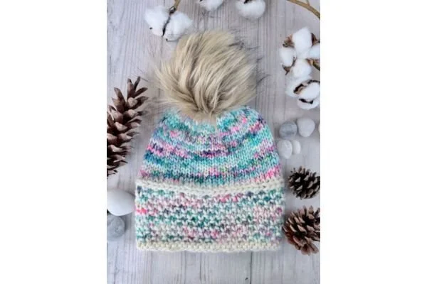 A hand knit hat with a brown pompom on a light wood table with pine cones