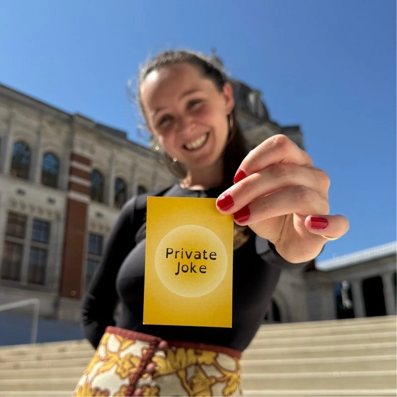 Olive stands smiling happily with one hand behind her back and the other foremost. In this hand she is holding a bright yellow Private Joke playing card for the viewer to see. Behind her is a scene from one of her many adventures around the world!