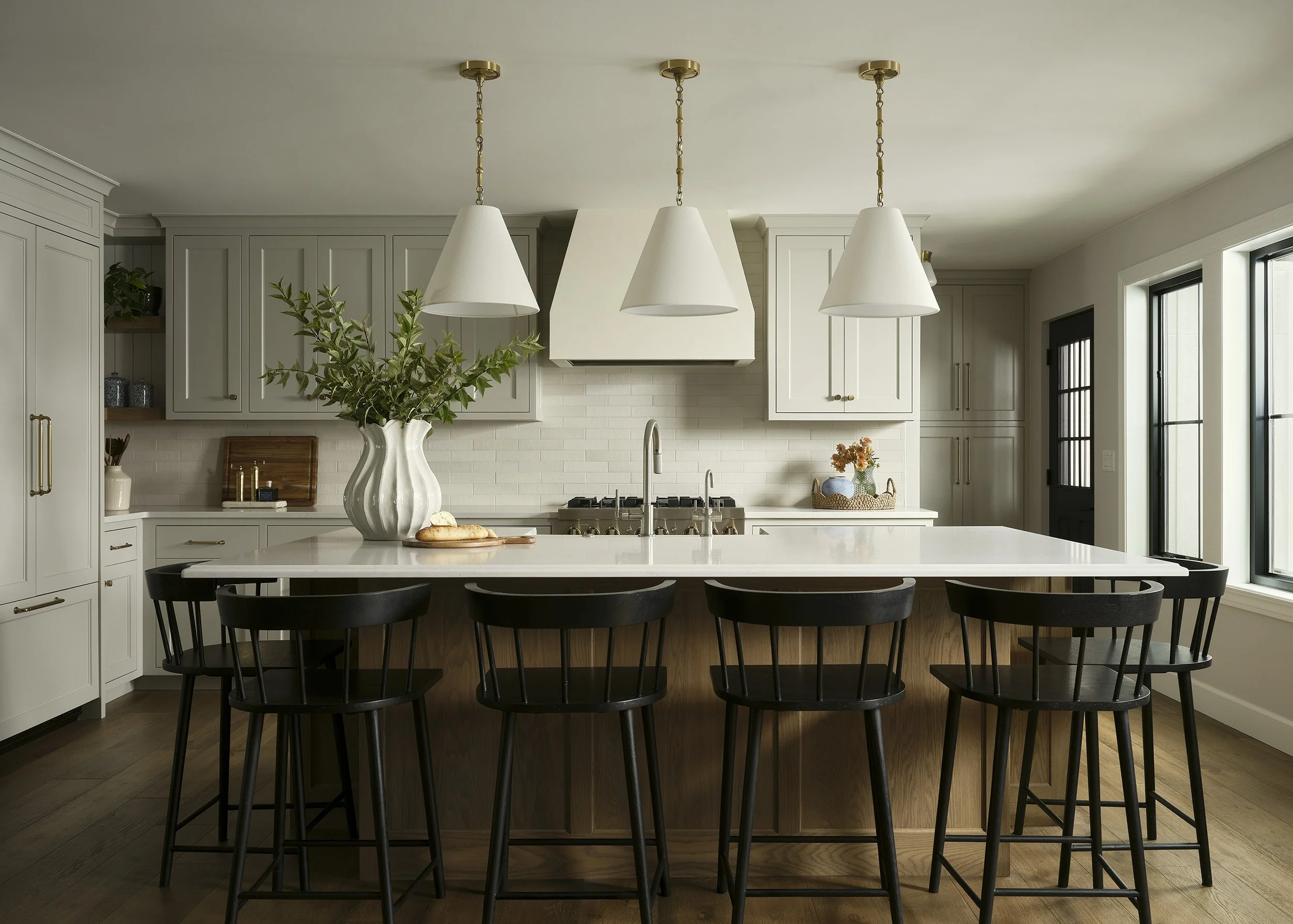 Modern kitchen with white cabinets, a large white island with black chairs, pendant lights, and a vase with greenery on the island.