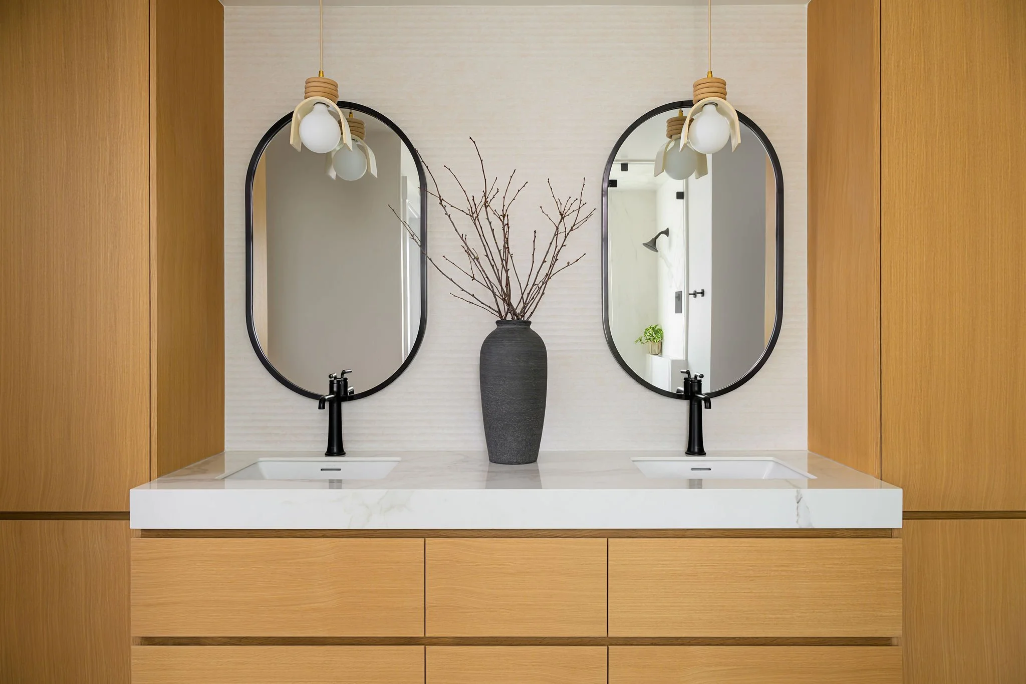 A modern bathroom double vanity with two oval mirrors, black faucets, and a large black vase with bare branches between the mirrors. Wooden cabinetry surrounds the vanity, and pendant lights hang above each mirror.
