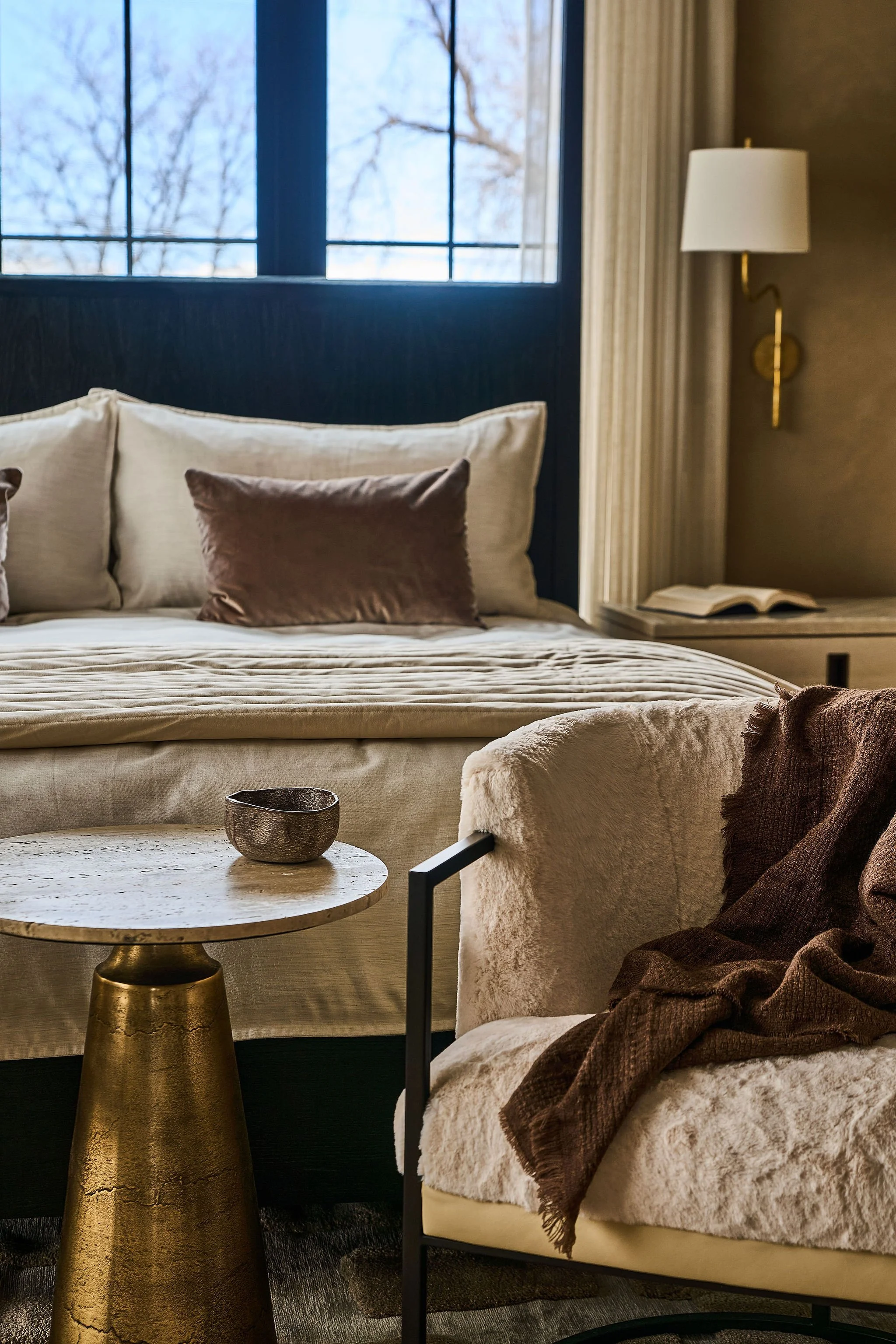 neutral primary bedroom with faux fur chair at foot of bed, brass wall sconces and sheer drape