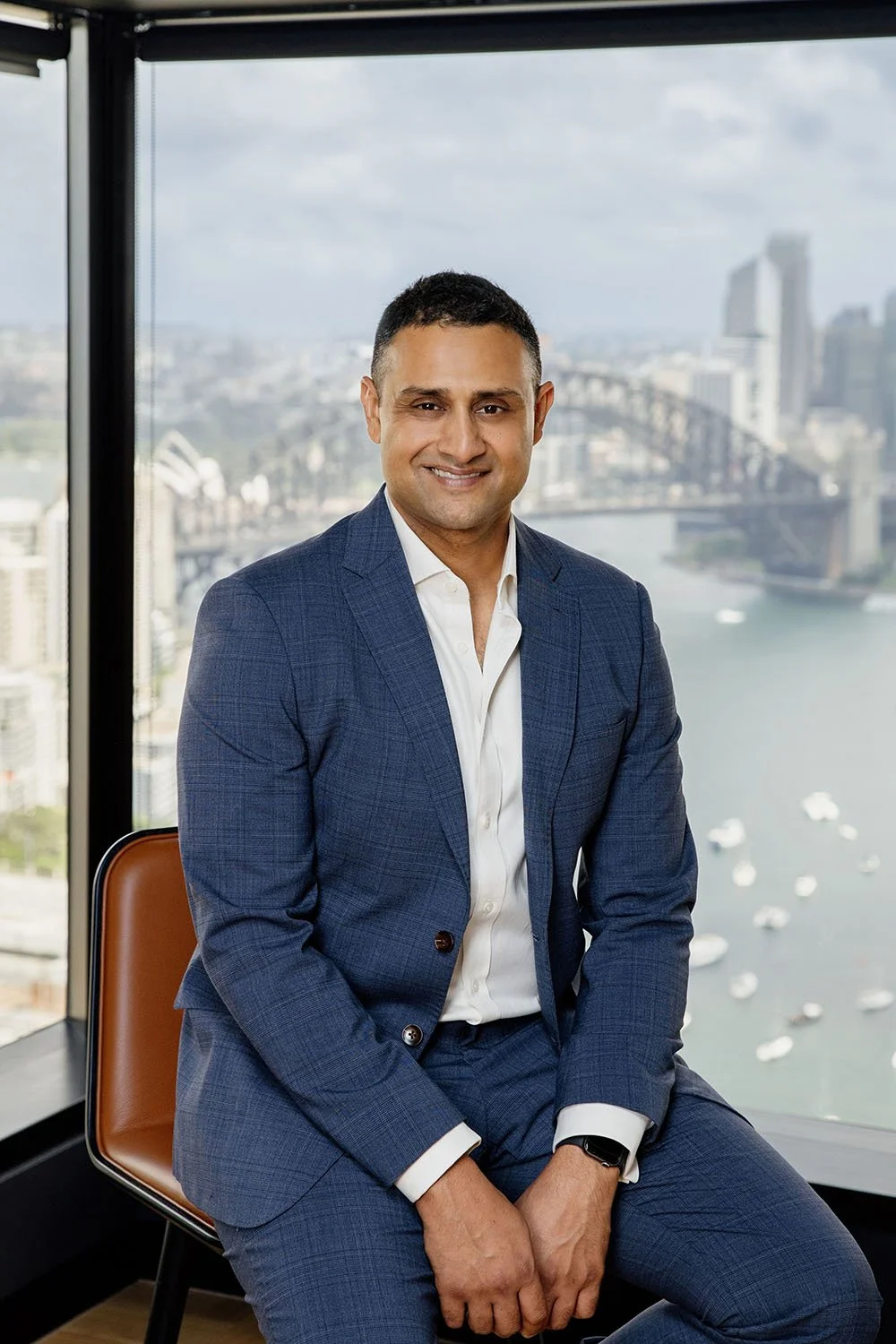 Simran Dhillon in a navy blue suit + white shirt, sitting in front of a large window overlooking Sydney Harbour and the Harbour Bridge.