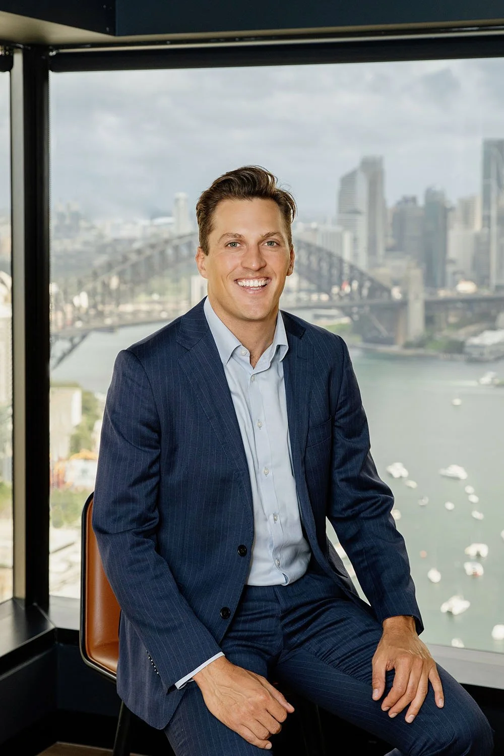 James Weyler in a navy blue suit with a white shirt, sitting in front of a large window overlooking Sydney Harbour and the Harbour Bridge.