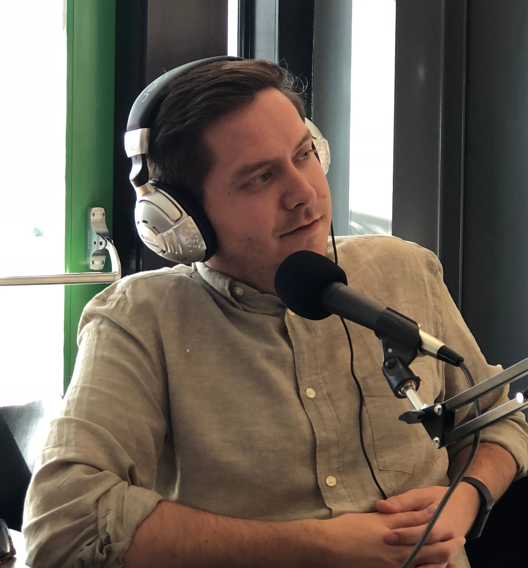 A man wearing headphones speaking into a microphone, sitting indoors near a window.