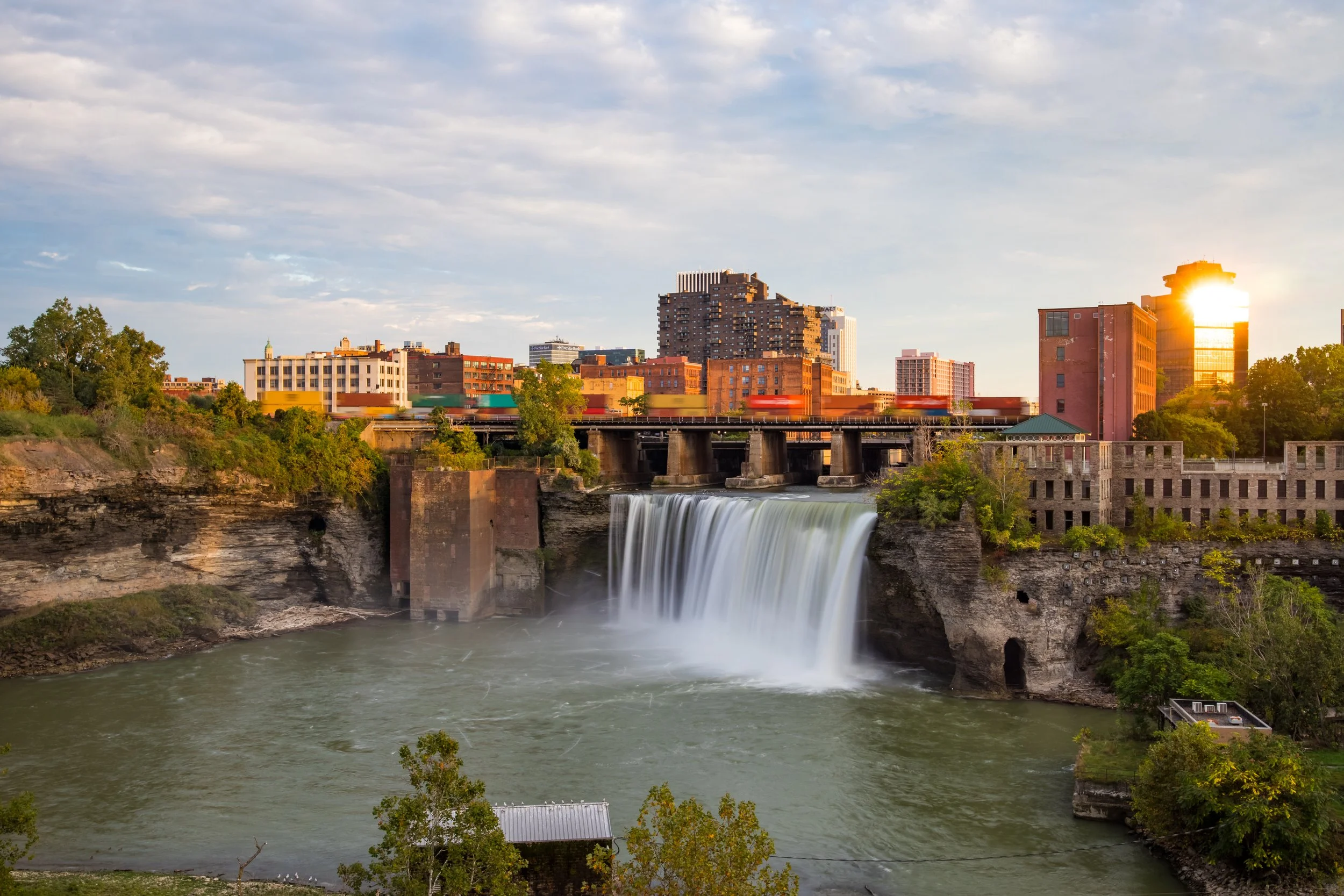 City skyline featuring buildings with the sun setting behind, a river waterfall in the foreground, and trees along the riverbank.