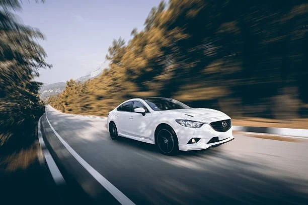 A white sedan car driving on a winding mountain road with blurred trees on both sides.