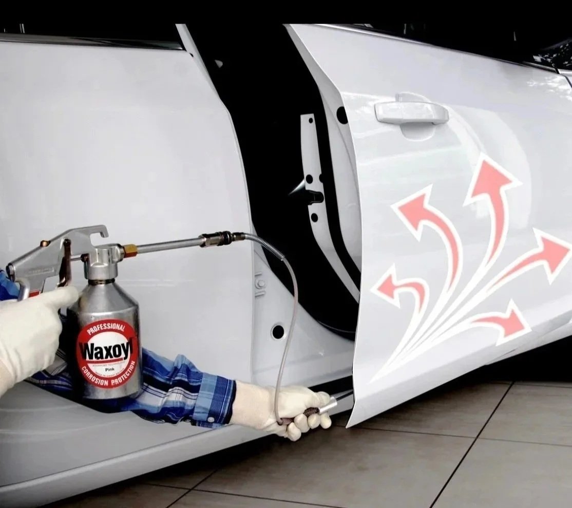 A person is spraying a protective coating inside a car door panel with a professional spray gun labeled 'Waxoyl'. The spray is directed onto the inner surface of the car door.