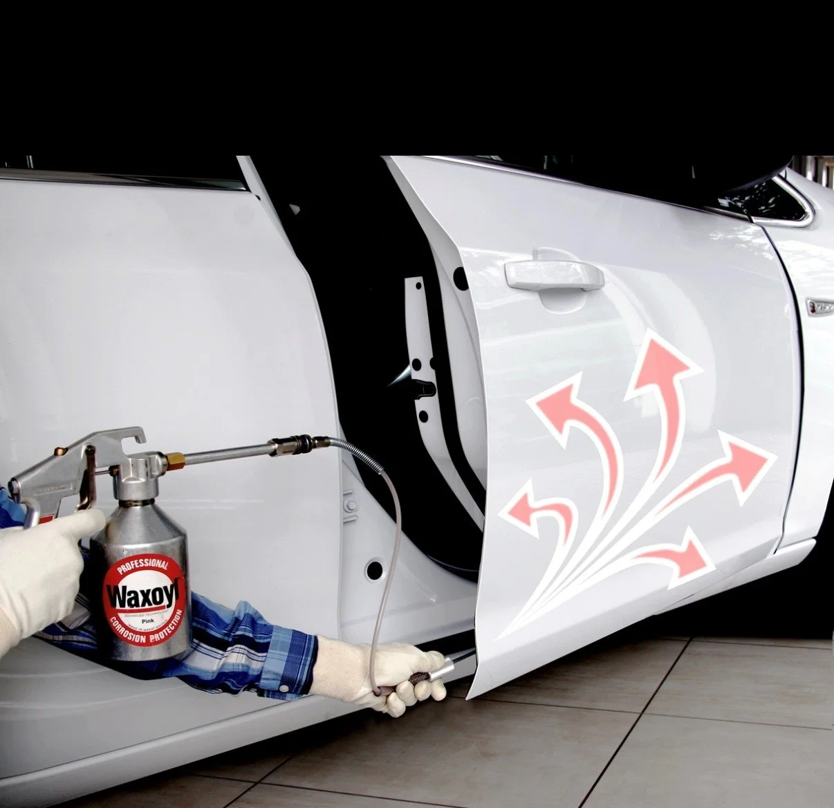 A person is spraying a protective coating inside a car door panel with a professional spray gun labeled 'Waxoyl'. The spray is directed onto the inner surface of the car door.