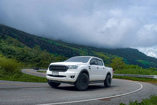 White pickup truck driving on a mountain road with green hills and cloudy sky in the background.