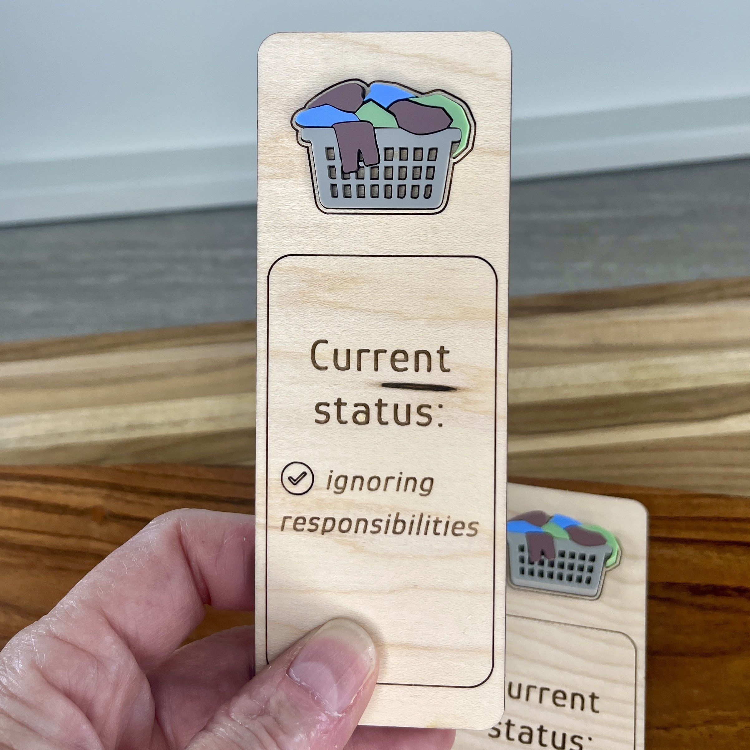 handheld maple bookmark featuring multi-coloured acrylic overflowing laundry basket embellishment engraved with the phrase "Current status: ignoring responsibilities".