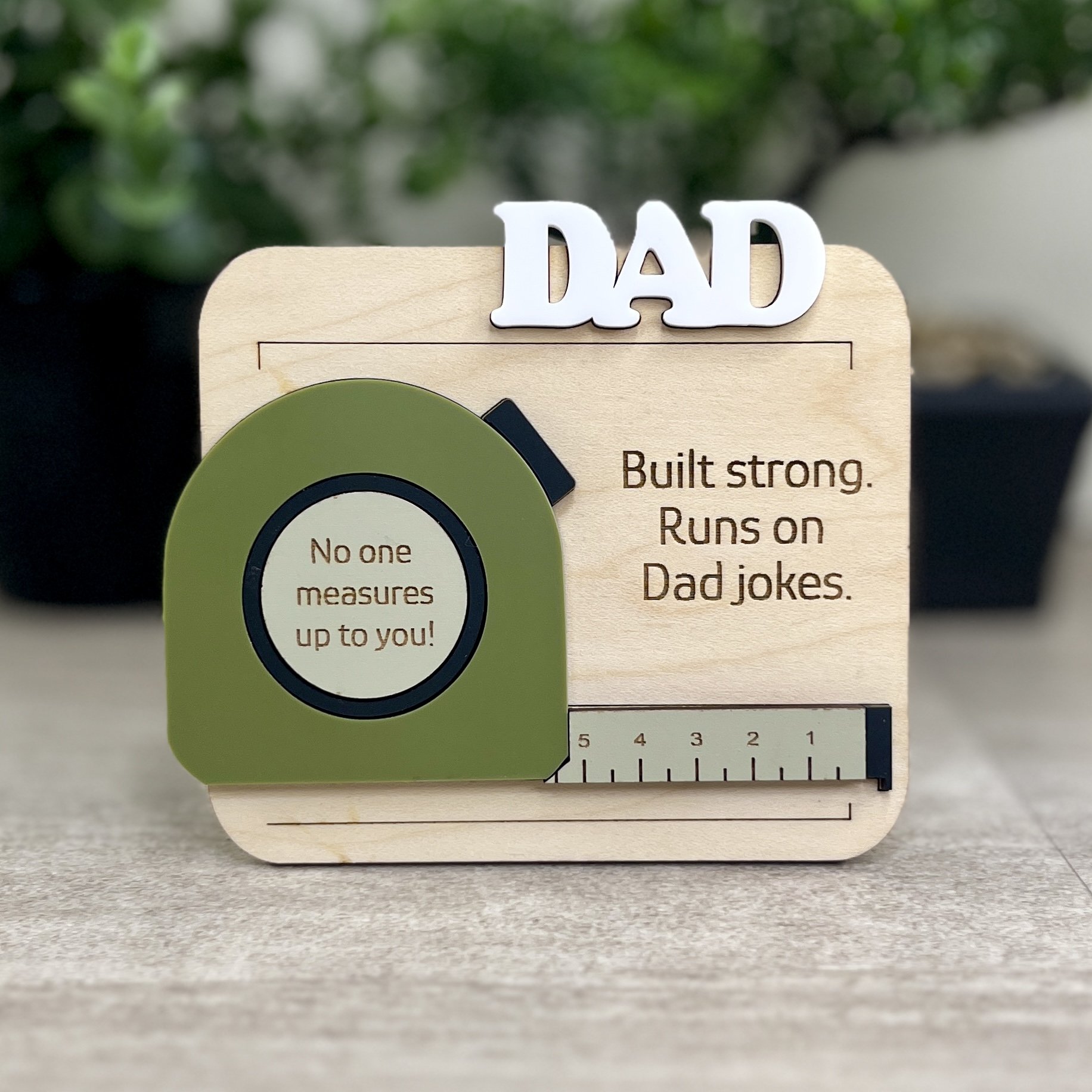 Front view of laser cut maple magnet with raised olive acrylic tape measure design. Engraved with quotes "Built strong.  Runs on Dad jokes." and "No one measures up to you".