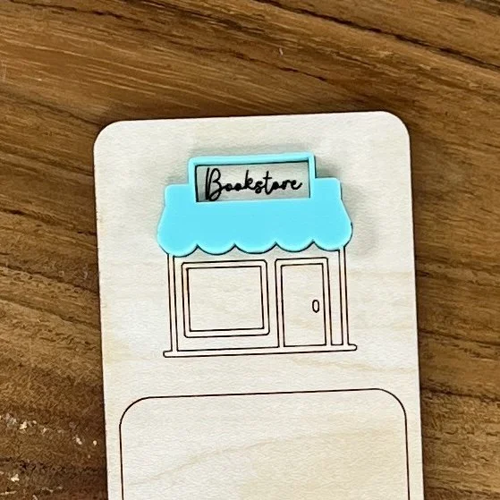 close up maple bookmark featuring mint acrylic bookstore awning above a bookstore storefront illustration engraved with the phrase "I brake for bookstores".
