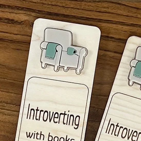 close up maple bookmark featuring a comfy sand coloured acrylic chair with sage blanket and coffee mug embellishment engraved with the phrase "Introverting with books".