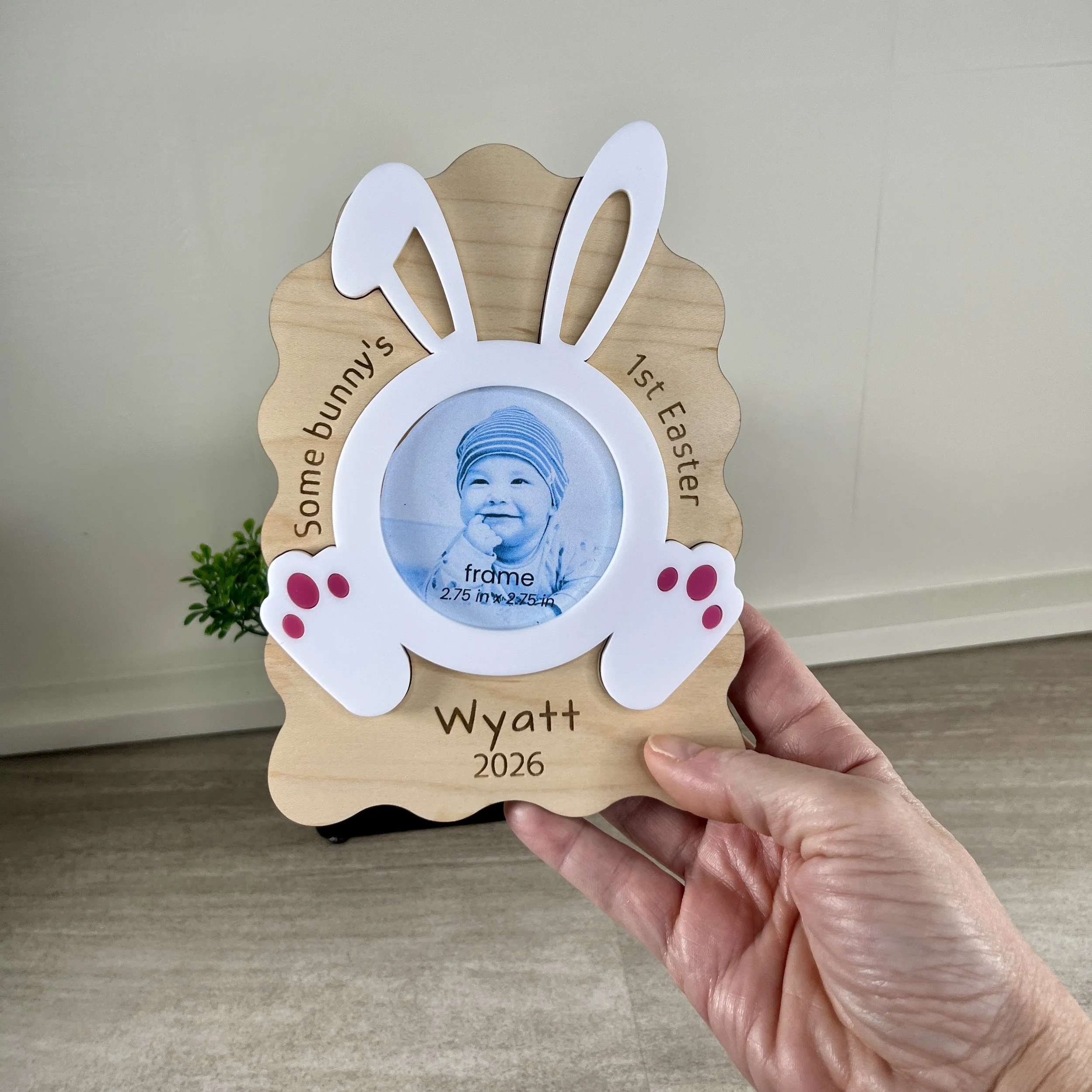 Personalized laser-cut maple wood Baby's First Easter photo frame with bunny ears and a circular opening for a baby's photo, held in hand for scale.