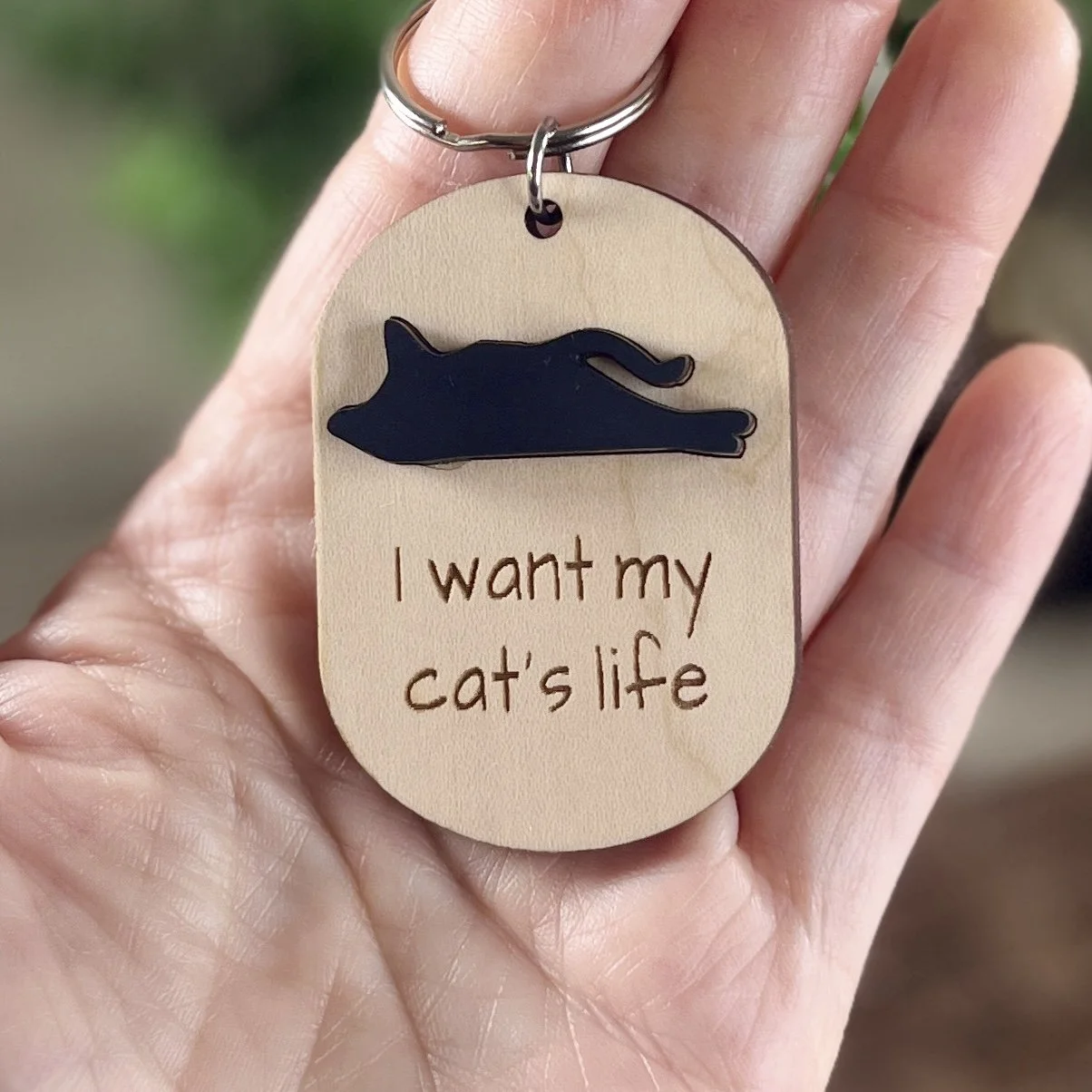 handheld view of laser cut maple acrylic keychain displaying silhouette of cat in matte black and engraved with the saying "I want my cat's life".