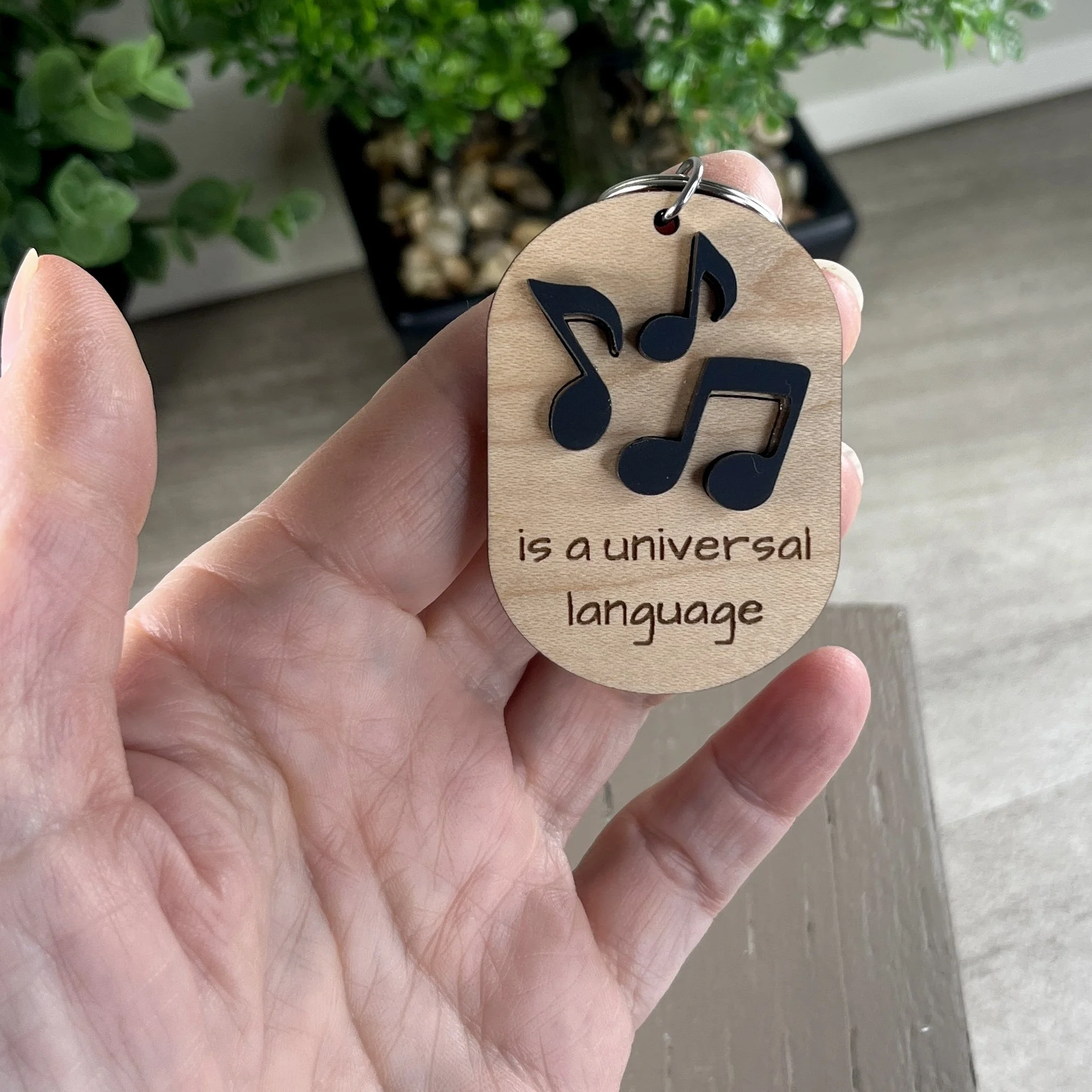 handheld laser cut maple wood keychain featuring three raised musical notes in matte black acrylic and the engraved phrase "is a universal language."