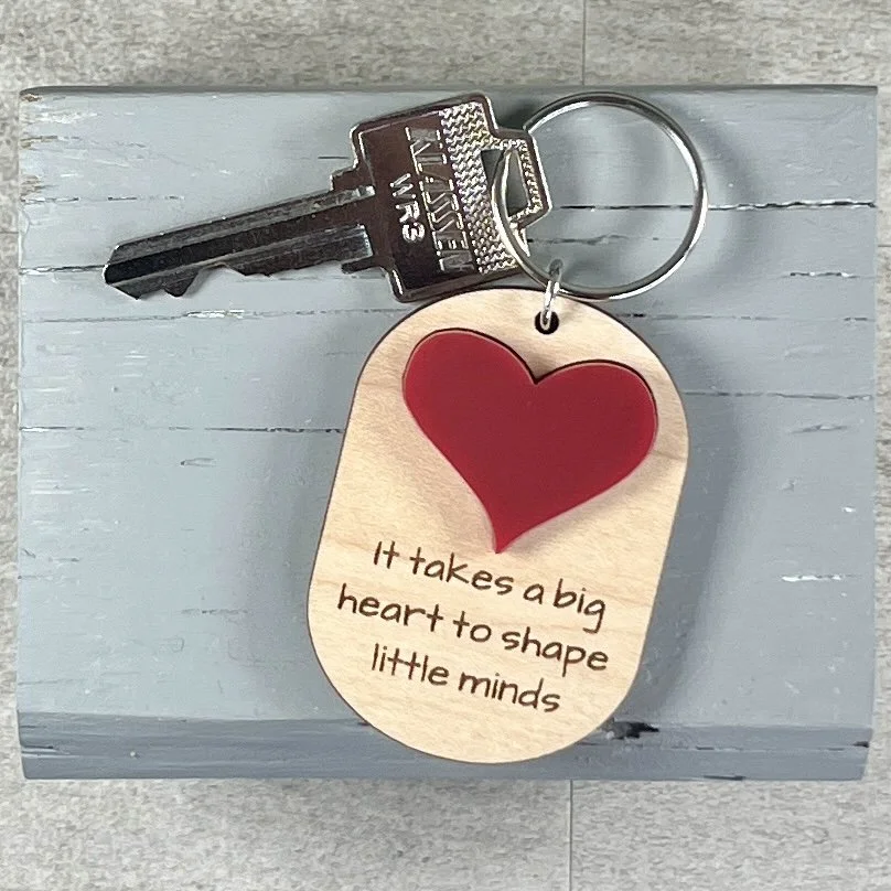 lifestyle shot of laser cut maple wood keychain featuring raised matte red acrylic heart and engraved with the phrase "It takes a big heart to shape little minds".