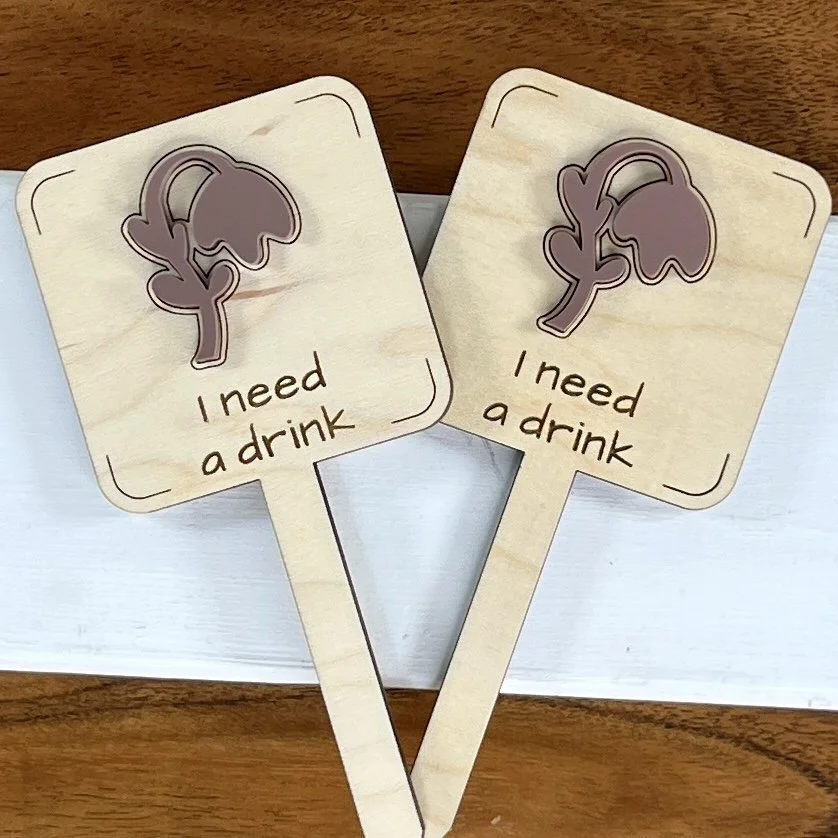flat lay maple plant stake featuring a droopy plant embellishment engraved with the phrase "I need a drink".