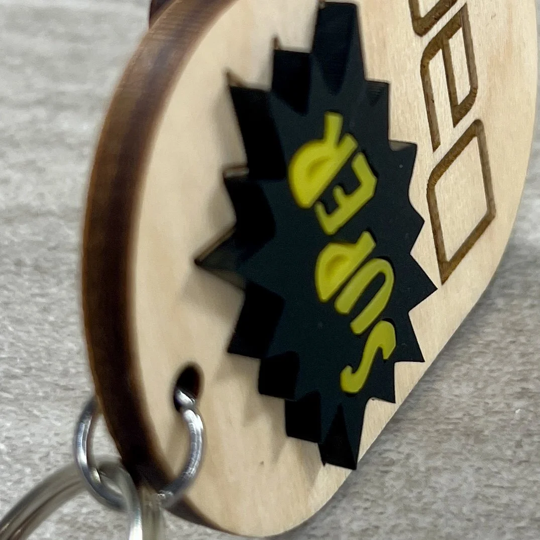 side view of wood keychain showing raised black and yellow comic book burst shape reading Super with Dad engraved beneath.