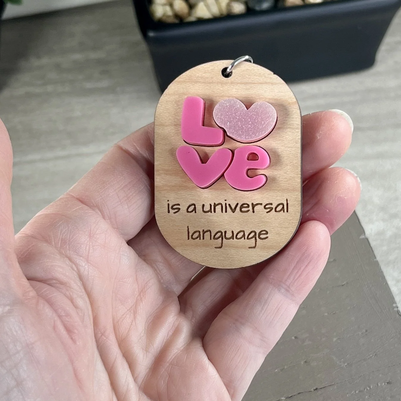 handheld laser cut maple wood keychain featuring layered pink acrylic letters spelling "Love" with heart details and the engraved phrase "is a universal language."