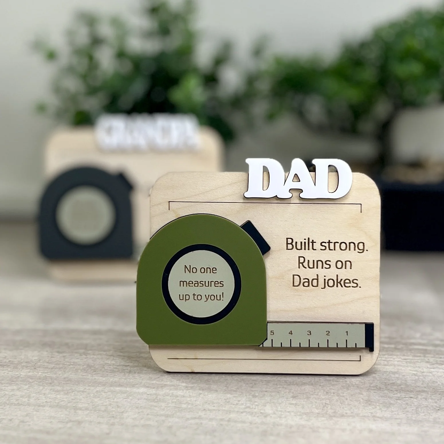 Set of laser cut maple magnet with raised acrylic tape measure design. Header features raised white text. Engraved with quotes "Built strong.  Runs on Dad jokes." and "No one measures up to you".
