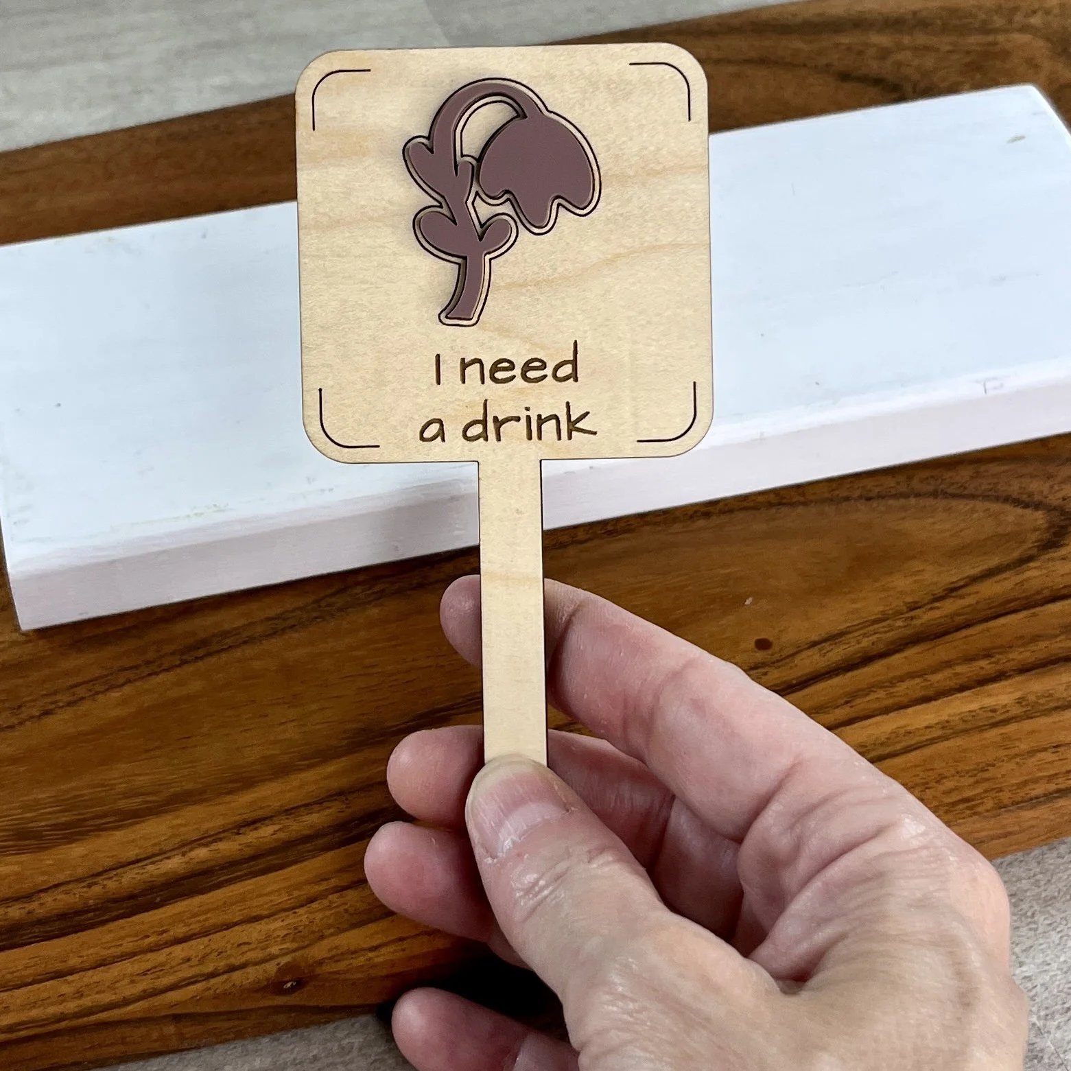 handheld maple plant stake featuring a droopy plant embellishment engraved with the phrase "I need a drink".