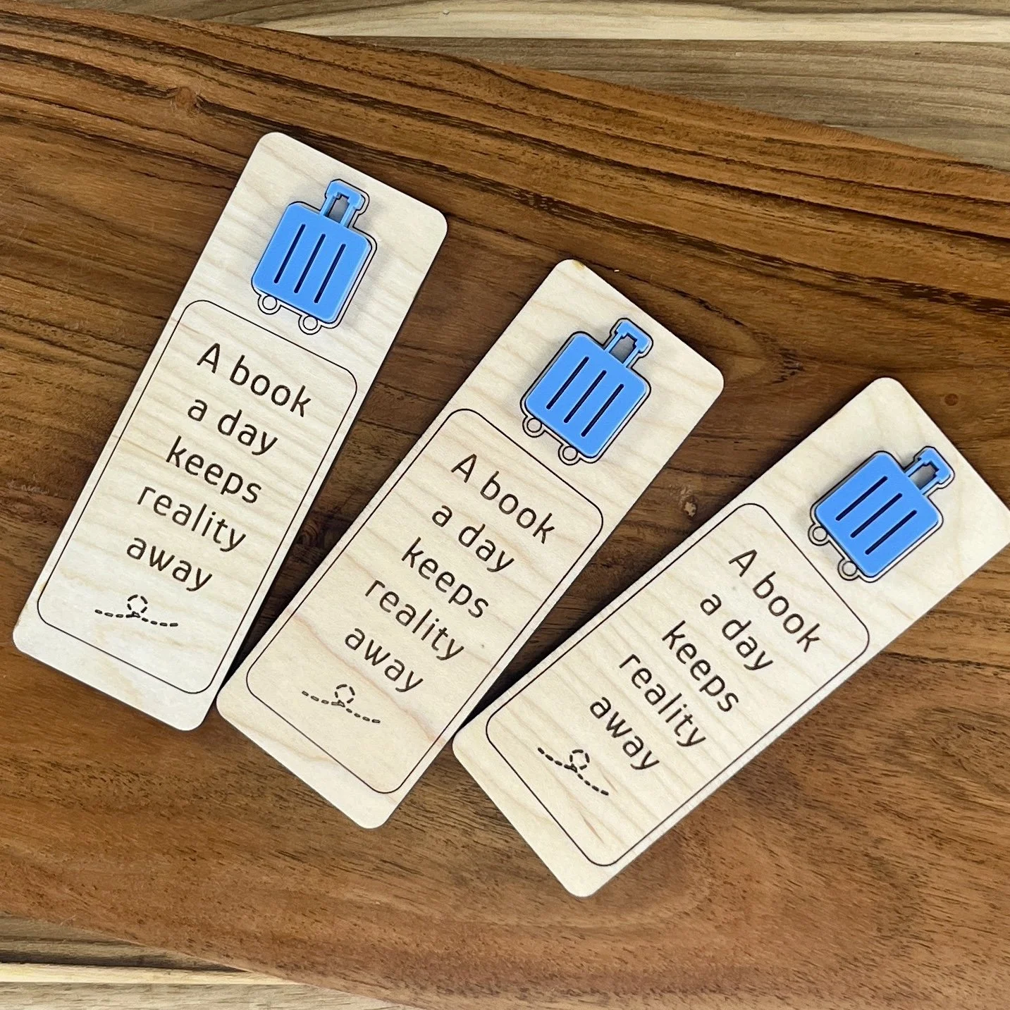 flat lay maple bookmark featuring blue acrylic suitcase embellishment engraved with the phrase "A book a day keeps reality away".